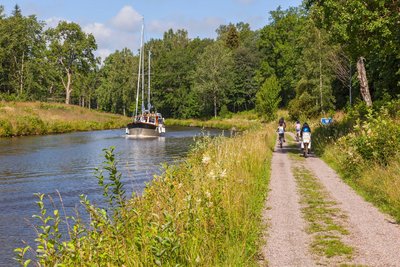 Rechts entlang eines Kanals verläuft ein gekiester Radweg. Im Hintergrund sind Radfahrer unterwegs, auf dem Kanal fährt ein Boot.
