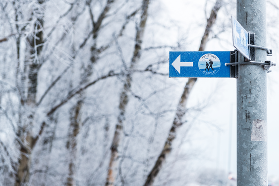 Wanderwegweiser des Küstenwanderwegs Kuststigen An einem Pfahl rechts im Bild ist ein blaues Schild, das auf den Wanderweg Kuststigen verweist, angebracht. Links des Pfahls erkennt man verschneite Bäume.