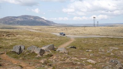Fahrt durch das Hochland am Uxahryggir-Pass Ein einzelnes Auto fährt auf einer Schotterstraße durch eine weite, kahle Hochlandlandschaft mit Bergen in der Ferne. Der Himmel ist bewölkt, die Vegetation spärlich.