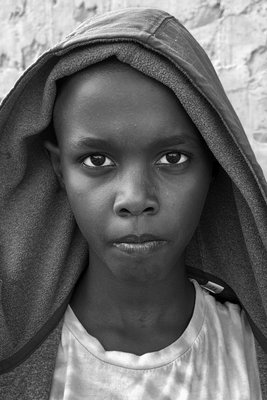 Nahsichtiges schwarz-weiß Porträt einer jungen Afrikanerin, die direkt in die Kamera schaut. Ihre Haare und Schultern werden von einem einfarbigen, dunklen Tuch oder einer Kapuze bedeckt.