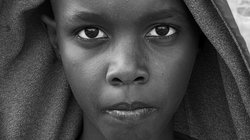 Nahsichtiges schwarz-weiß Porträt einer jungen Afrikanerin, die direkt in die Kamera schaut. Ihre Haare und Schultern werden von einem einfarbigen, dunklen Tuch oder einer Kapuze bedeckt.