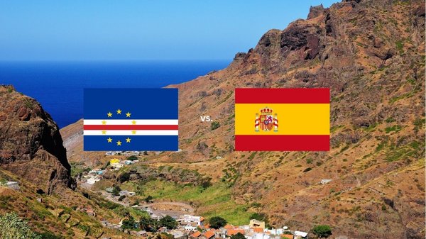 Auf dem Bild sind die Flaggen der Kapverden und von Spanien abgebildet. Im Hintergrund ist eine bergige Landschaft der Kapverden direkt am Meer abgebildet.