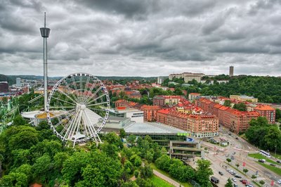 Links im Vordergrund erhebt sich zwischen Bäumen eine Riesenrad und weitere Fahrgeschäfte, rechts im Bild blickt man auf Göteborg, darüber dräut ein grauer, wolkenverhangener Himmel.