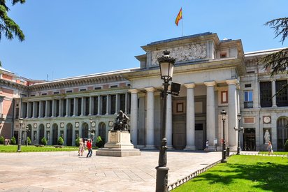 Man blickt auf die Fassade des Prados, rechts im Bild ist das Säulenportal mit dem Eingang zum Gebäude. Links setzt sich der Seitenflügel bis zu seiner Ecke fort, während der rechte Flügel nur noch ein kurzes Stück im Bild zu sehen ist. Auf dem Platz vor dem Eingangsportal steht ein Denkmal, davor zwei Menschen, die fotografieren. Der Himmel ist leuchtend blau.