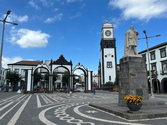 Rechts im Bild steht auf einem Sockel die Statue eines Mannes. Der Platz, auf dem sich die Statue befindet, wir dem Hintergrund von einem dreibögigen Tor, einem Uhrturm und Gebäuden begrenzt. Die Architektur ist weiß mit schwarzen Einfassungen. Am blauen Himmel darüber schweben einzelne weiße Wolken.
