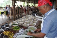 Eine Reisegruppe erwartet am Tisch die Ergebnisse des Kochkurses in der Roça São João auf São Tomé, während der Koch die Teller anrichtet.