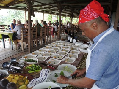 Eine Reisegruppe erwartet am Tisch die Ergebnisse des Kochkurses in der Roça São João auf São Tomé, während der Koch die Teller anrichtet.