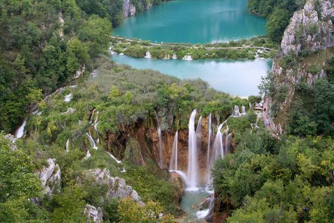 Ein türkisblauer See im Plitvicer Nationalpark, Kroatien, umgeben von dichten, grünen Wäldern. Das klare Wasser spiegelt die Bäume wider, während kleine Wasserfälle in den See fließen. Ein Holzsteg führt durch die Landschaft.