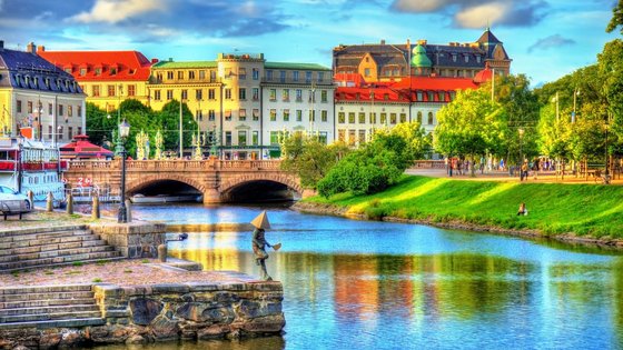 Stadt am Wasser: Göteborg Man schaut über einen Flusslauf auf eine Brücke und historische Häuserzeile im Hintergrund, vorne am Wasser steht eine kleine Skulptur eines Menschen mit einer Pyramide auf dem Kopf.