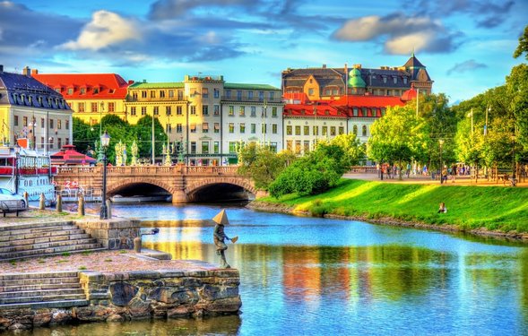 Man schaut über einen Flusslauf auf eine Brücke und historische Häuserzeile im Hintergrund, vorne am Wasser steht eine kleine Skulptur eines Menschen mit einer Pyramide auf dem Kopf.