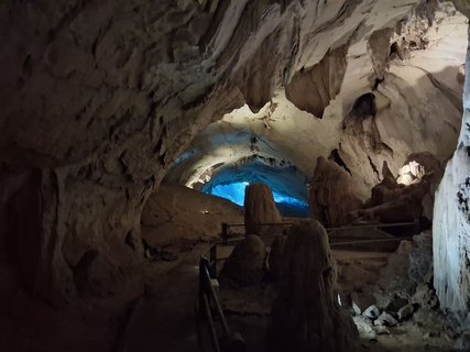 Das Bild zeigt das Innere einer Höhle, die mit Wegen und Handläufen erschlossen ist. In der Bildmitte sieht man ein Oval leuchtend blauen Lichts.