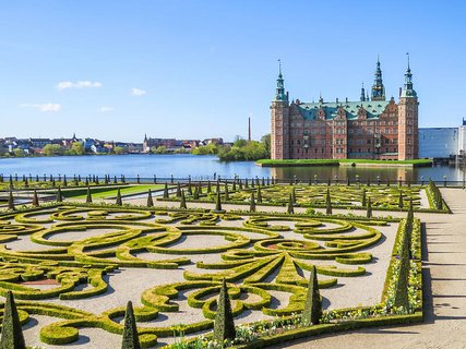 Ein von Wasser umgebenes Schloss. Davor befindet sich ein ordentlich hergerichteter, verschnörkelter Garten.