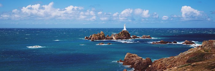 Der Corbière-Leuchtturm auf der Kanalinsel Jersey, umgeben von einer felsigen Küstenlandschaft bei Ebbe, mit einem Fußweg, der durch das Wasser zum Leuchtturm führt.