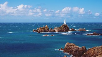 Der Corbière-Leuchtturm auf der Kanalinsel Jersey, umgeben von einer felsigen Küstenlandschaft bei Ebbe, mit einem Fußweg, der durch das Wasser zum Leuchtturm führt.