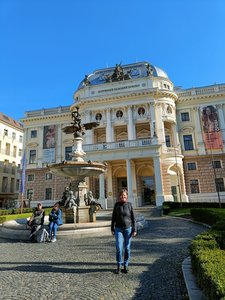 In der Bildmitte erhebt sich das historistische Slowakische Nationaltheater mit seiner weiß-gelben reich verzierten Fassade. Auf dem Platz davor befindet sich ein rundes Wasserbassin, dessen Wasserspeier in der Mitte eine Ganymed-Szene darstellt. Das pbere Bilddrittel wird von blauem Himmel ausgefüllt.