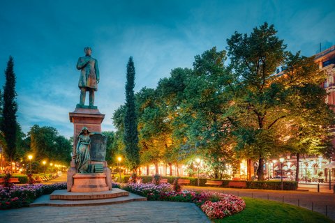 Links im Vordergrund steht die Statue eines Mannes auf einem Sockel im Blumen- und Baum-bestandenen Esplanadi-Park von Helsinki. Wegen der Abenddämmerung sind die Schaufenster der Geschäfte an der Seite des Parks beleuchtet.