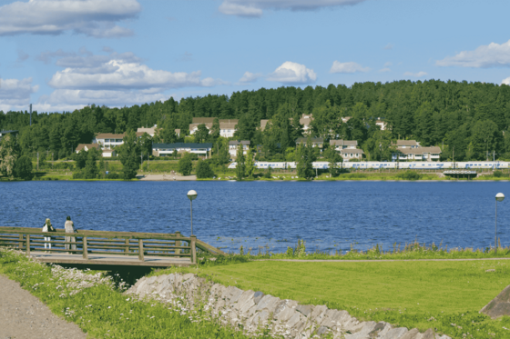 In der Bildmitte verläuft quer ein Fluss. Im Vordergrund mündet ein kleiner Zufluss in ihn, der von einer Brücke überfangen ist, auf der zwei weibliche Personen stehen. Im Hintergrund fährt ein Zug entlang des Flusses, dahinter erhebt sich ein Hügel, auf dem Häuser stehen und der dicht mit Bäumen bewachsen ist. 