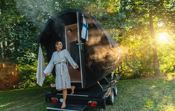 Mobile Sauna in Finnland Zwei Frauen steigen die Treppen eines Saunawagens hinab, der vor eine paar Bäumen steht, durch die das Sonnenlicht scheint.