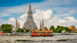Über eine Wasserfläche auf der sich zentral ein traditionelles und links im Bild zwei moderne Langboote befinden, blickt man auf auf einen hohen weißen Tempel mit verschiedenen Goldelementen, den Wat Arun in Bangkok.