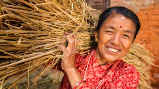 Eine einheimische, älter Frau trägt Stroh auf den Schultern und lächelt in die Kamera
