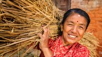 Eine einheimische, älter Frau trägt Stroh auf den Schultern und lächelt in die Kamera