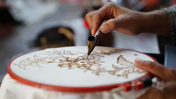 Eine Nahaufnahme zeigt eine Hand, die mit einem traditionellen Tjanting-Werkzeug feine Muster auf einen Batik-Stoff aufträgt.