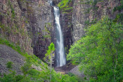 Ein Wasserfall ergießt sich zwischen Felswänden in die Tiefe, vorne stehen Bäume.