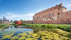 Im Vordergrund befindet sich eine mit Entengrütze bedeckte Wasserfläche, rechts dahinter erstreckt sich in die Bildtiefe hinein ein trutziges Gebäude.