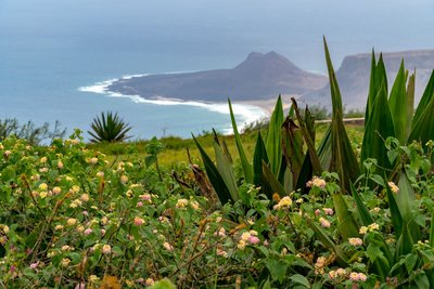 Der Blick vom Monte Verde über  São Vicente gehört zu den Sehenswürdigkeiten der Kapverden, die man sich nicht entgehen lassen sollte. Der Vordergrund des Fotos wird von grüner Vegetation eingenommen, dahinter in der Tiefe sieht man den blauen Ozean mit einer Halbinsel.