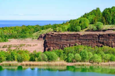 Am vorderen Bildrand zieht sich ein Wasserstreifen entlang, in dem sich der Himmel und der Uferbewuchs bestehend aus Schilf und Laubbäumen spiegelt. Rechts in der Bildmitte befindet sich eine Steilwand, links davon eine unbewachsene, hellrotbräunliche Fläche. Hinter ihr und der Steilwand schließt sich eine mit Bäumen bewachsene, nach rechts ansteigende Hügellandschaft an. Dahinter links im Bild erstreckt sich eine Wasserfläche bis zum Horizont, wo sie auf blauen Himmel trifft.
