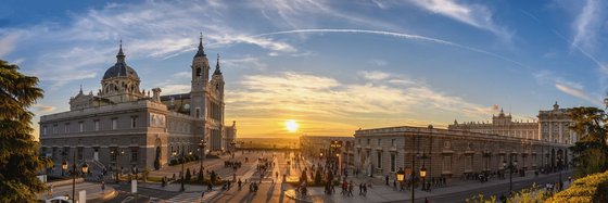 Das überbreite Bild zeigt ein Architektur-Panorama von Madrid. Auf der linken Seite des Bildes befindet sich die Almudena-Kathedrale, rechts der Königspalast. Zwischen den beiden Gebäuden scheint die tiefstehende, diffuse Sonne. Der Himmel ist blau mit weißen Wolken und Chem-Trails.