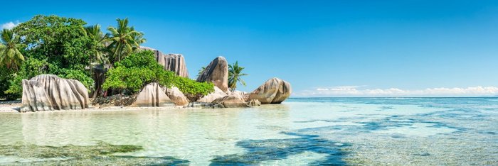 Ein Strand mit hellen Granitfelsen, klarem Wasser und flachem Ufer. Im Hintergrund befinden sich dichte grüne Bäume und Pflanzen."
