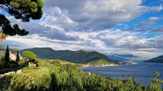 Die linke Bildhälfte wird von grün bewachsenen Hängen und im Hintergrund höheren Hügeln dominiert, während die rechte untere Bildhälfte von einer dunkelblauen Wasserfläche eingenommen wird. Im Bildmittelgrund setzen sich die grünen Berge fort. Der Himmel darüber ist mit großen hellgrauen Wolken verhangen.