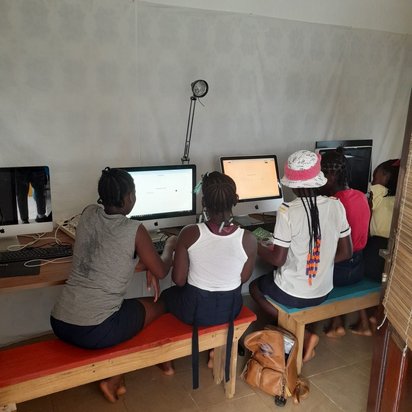 Man sieht eine Reihe von afrikanischen Jugendlichen von hinten, die auf einer Bank sitzen und auf Computerbildschirme vor ihnen auf einem langen Tisch schauen.