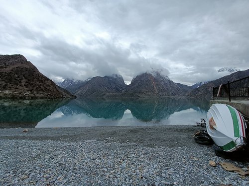 Der Iskandarkul-See in Tadschikistan Über ein kiesiges Ufer schaut man auf einen See, hinter dem sich Berge und ein grauer Himmel erheben. Rechts am Bildrand liegt ein kleines Boot kielaufwärts.