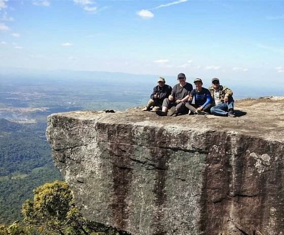 Ein flacher Felsen ragt von rechts in das Bild hinein, darauf sitzen vier Männer und schauen in Richtung der Kamera. Neben und hinter dem Felsen geht es tief hinab und man schaut weit ins Land, darüber diesig-blauer Himmel.