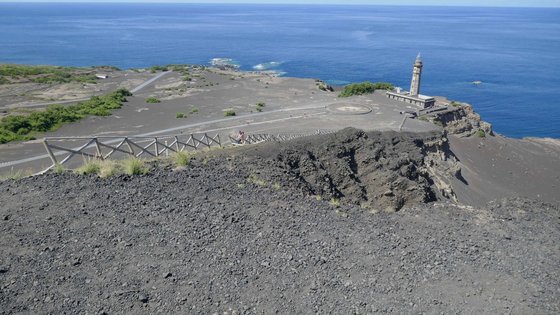 Vom Bildvordergrund bis über den Bildmittelgrund hinaus erstreckt sich eine graue Lavalandschaft bis an das blaue Meer im hinteren Bilddrittel. Rechts im Bildhintergrund steht ein kleiner, grauer Leuchtturm auf eine Erhebung über dem Meer.