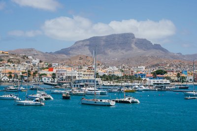 Mindelo mit seiner Musikszene gehört zu den wichtigsten Sehenswürdigkeiten der Kapverden. Das Foto zeigt die Stadt vom Wasser aus gesehen. Auf dem blauen Meer dümpeln weiße Yachten, hinter der Stadt erhebt sich ein karges Gebirge, darüber blauer Himmel.