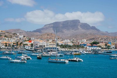Mindelo mit seiner Musikszene gehört zu den wichtigsten Sehenswürdigkeiten der Kapverden. Das Foto zeigt die Stadt vom Wasser aus gesehen. Auf dem blauen Meer dümpeln weiße Yachten, hinter der Stadt erhebt sich ein karges Gebirge, darüber blauer Himmel.