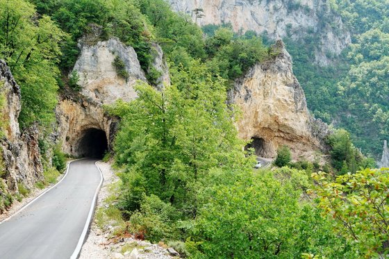 Links im Bild führt eine Straße in einen in den Fels geschlagenen Tunnel, rechts im Bild sieht man, dass die Straße wieder aus dem Berg hinausführt. Die umgebenden grau-braunen Felsen sind mit vielen Bäumen bewachsen.