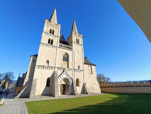 Es erhebt sich eine eher gedrungen wirkende, grau-beige verputzte Kirche, deren Fassade von zwei Türmen geziert wird. Das Bild ist durch einen Filter mit übertriebener Froschperspektive verzerrt, so dass alle Bauelemente zur Bildmitte streben. Der Bildraum vor der Kirche wird durch eine Wiese ausgefüllt, um die Kirche herum blauer Himmel.