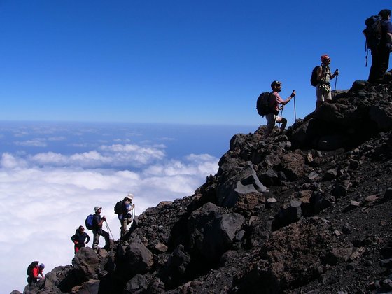 Das Bild wird von links unten nach rechts oben durch einen Berggrat geteilt, auf dem eine Gruppe von Wanderern hintereinander dem Gipfel entgegenstreben. Links im unteren Bereich sieht man eine weiße Wolkendecke, das obere Drittel des Bildes wird von blauem Himmel eingenommen.