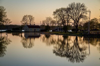 Die untere Bildhälfte wird von einer spiegelnden Wasserfläche eingenommen, die die dahinterliegenden Bäume sowie ein gelbes Haus reflektiert.
