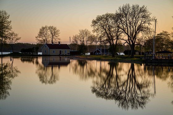 Sjötorp am westlichen Ende des Göta-Kanals Die untere Bildhälfte wird von einer spiegelnden Wasserfläche eingenommen, die die dahinterliegenden Bäume sowie ein gelbes Haus reflektiert.