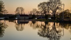 Die untere Bildhälfte wird von einer spiegelnden Wasserfläche eingenommen, die die dahinterliegenden Bäume sowie ein gelbes Haus reflektiert.