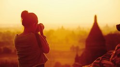 Auf Fotoreise im faszinierenden Asien Links im Bild steht eine Frau mit dem Rücken zum Betrachter. Sie blickt durch eine Kamera auf eine Tempelpagode rechts im Bildmittelgrund. Das gesamte Foto ist in orange-goldenes, aber dämmriges Sonnenlicht getaucht.