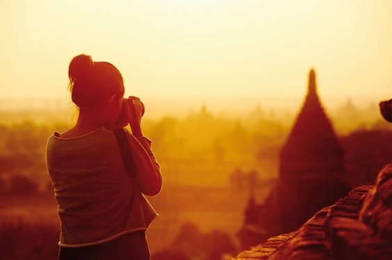 Auf Fotoreise im faszinierenden Asien Links im Bild steht eine Frau mit dem Rücken zum Betrachter. Sie blickt durch eine Kamera auf eine Tempelpagode rechts im Bildmittelgrund. Das gesamte Foto ist in orange-goldenes, aber dämmriges Sonnenlicht getaucht.