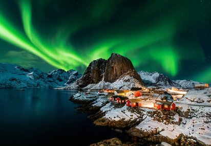 In Norwegen das magische Nordlicht erleben Über einer verschneiten Landschaft mit kleinen Häuschen erleuchtet das Nordlicht den Nachthimmel mit grünen Farbspielen.