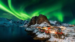 Über einer verschneiten Landschaft mit kleinen Häuschen erleuchtet das Nordlicht den Nachthimmel mit grünen Farbspielen.