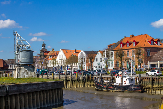 Der Hafen von Tönning Die untere Bildhälfte wird von einer Häuserzeile mit weiß getünchten Häusern, die rote Dächer haben, eingenommen, davor sieht man eine Hafeneinfahrt. Die obere Bildhälfte besteht aus klar blauem Himmel.